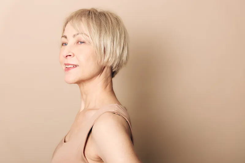 Smiling mature woman with short blonde hair wearing a beige top stands confidently against a soft beige background. Concept for aging beauty, confidence, and natural look.