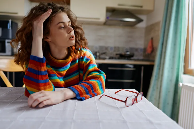 A young female in a colorful striped sweater sits at a kitchen table, resting her head on her hand with glasses nearby, conveying contemplation in a bright home setting.