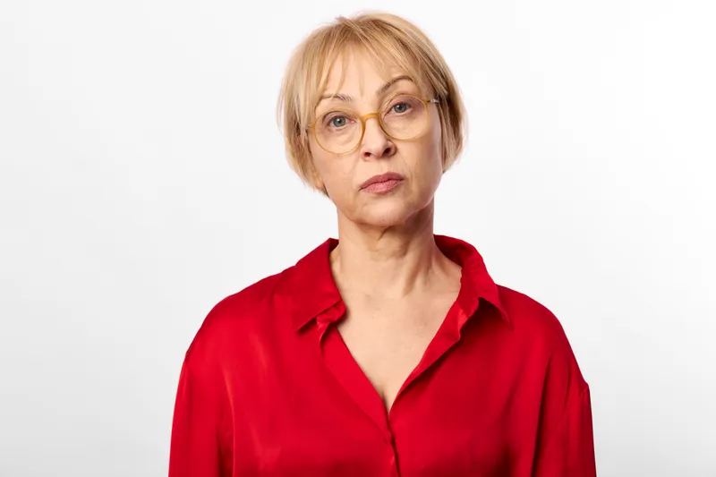 Woman portrait, older senior, blonde glasses, red shirt, studio background, calm serious face, neutral expression, adult female closeup headshot, soft light, wellness identity, minimalism