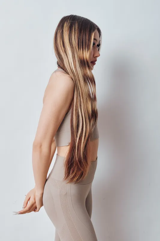 Young woman with long braided hair wearing beige outfit standing sideways against white background. Profile portrait with natural light and casual style.