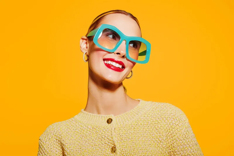 Smiling woman with large blue glasses and red lipstick poses against bright yellow background. She wears a textured yellow sweater and gold earrings. Concept of fashion, style, and vibrant colors.