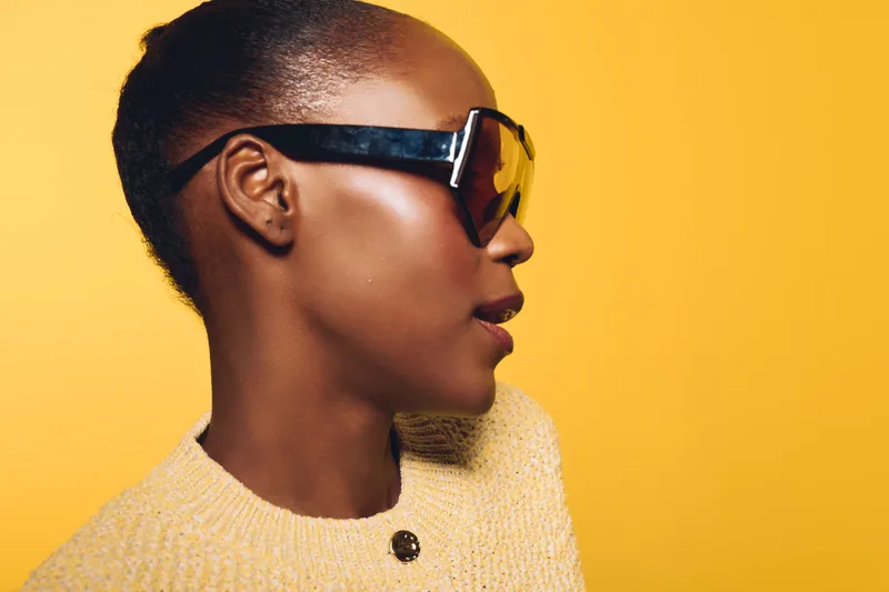 Profile of a stylish African woman with dark skin wearing oversized sunglasses and a beige knitted sweater. The bright yellow background emphasizes modern fashion and confident attitude.