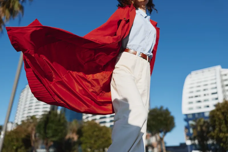 Cape superhero fashion in street setting, wind lifts cape in outdoor city scene, confident model poses against blue sky and buildings, dynamic motion and modern style