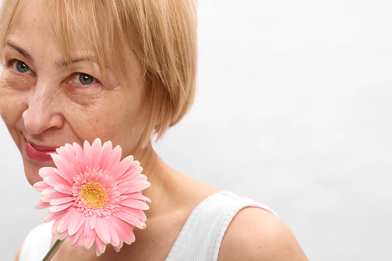 Elderly woman with blonde hair holds a pink gerbera flower close to her cheek, soft daylight on a white background, beauty and aging concept, portrait, calm tenderness