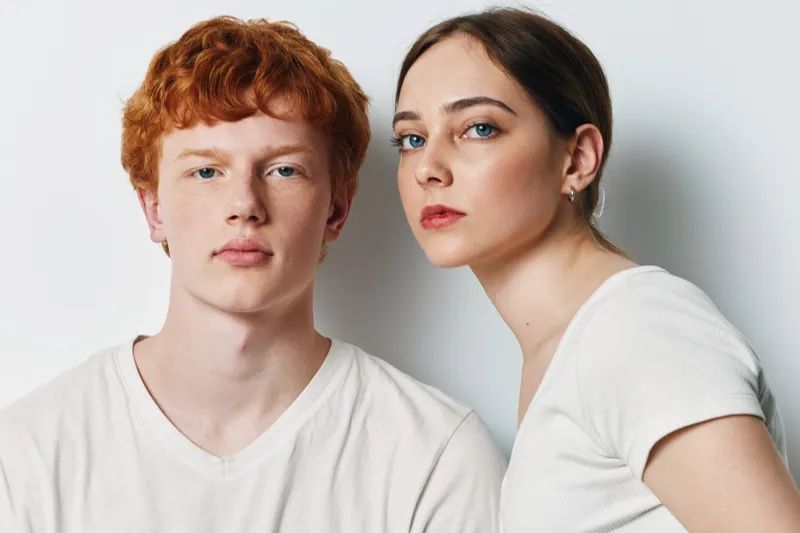 Portrait of young man and woman with serious expressions in white shirts against plain background. Closeup of teenagers with natural skin and casual hairstyle posing indoors.