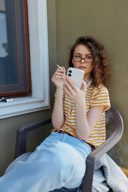 Casual woman with striped shirt and glasses sits on a chair outside a home, using a smartphone. Relaxed lifestyle, everyday moment, natural light, outdoor setting.