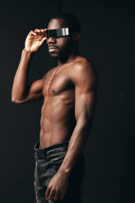 African man shirtless with futuristic glasses in creative conceptual visual style, posing against dark background, showcasing modern fashion and confident expression in studio lighting.