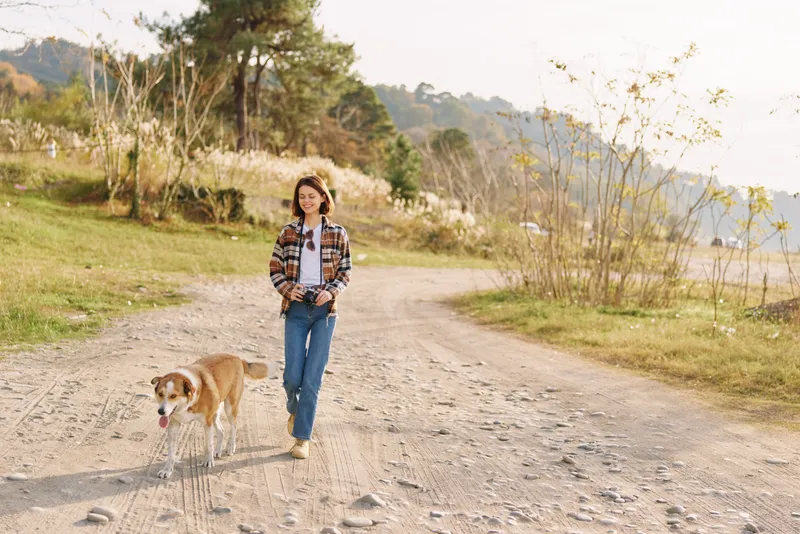 Woman walking a dog along a gravel path in a park, casual outfit, open space, trees in the background, peaceful outdoor moment, sunshine and natural scenery for lifestyle and leisure.