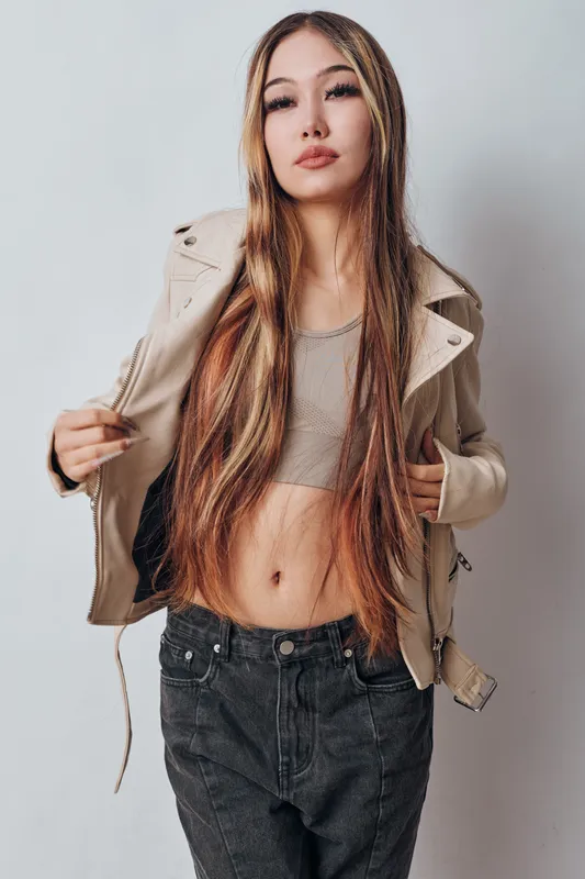 Young woman with long hair, casual outfit and confident pose standing against neutral background. Fashionable female in crop top, jacket and jeans showing style and attitude.