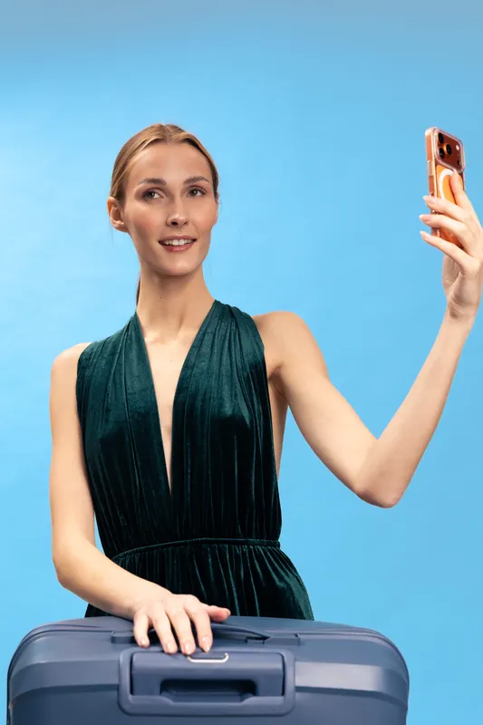 Young woman wearing elegant dress takes selfie with smartphone while standing behind suitcase on blue background. Concept for travel, vacation, lifestyle, and digital communication.