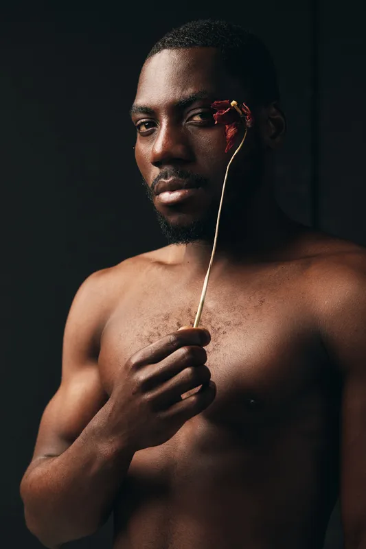 Creative conceptual portrait of African man with bare chest holding dried flower near face in dark studio. Artistic visual style with moody lighting and thoughtful expression on model.