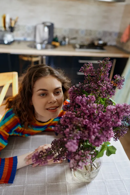 Woman rests at a table indoors, smiling, admiring a purple lilac bouquet in a glass vase, home setting, cozy kitchen in background, bright casual lifestyle moment, warm light, intimate still life