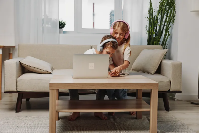 Two children wearing headphones interact happily while using a laptop on a wooden table in a cozy living room with natural light and modern decor. Concept of learning and technology.