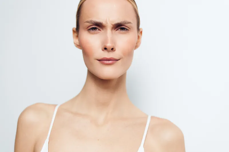 Young woman with serious expression, furrowed brows, looking directly at camera, neutral background, minimal makeup, natural lighting, closeup portrait, contemplative mood