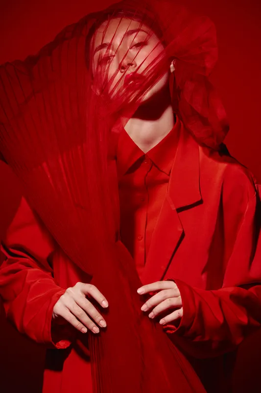 Young woman dressed in red holds translucent fabric in front of her face creating striking beauty and fashion concept with dramatic lighting studio shot. Creative elegance theme.