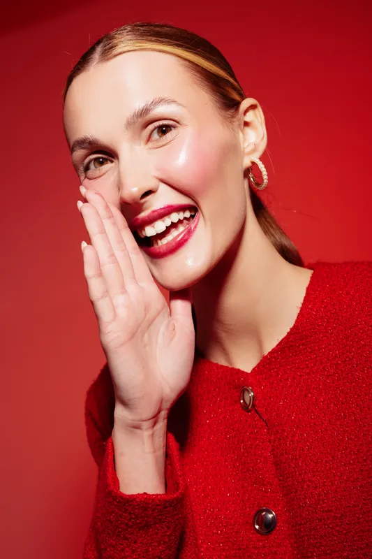 Smiling woman with shiny skin and red lipstick whispers with hand near mouth. She wears red clothing and hoop earrings on red background. Concept of communication and beauty.