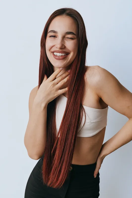 Young woman with long red hair smiling and winking while posing with hand on neck and hip against plain light background in casual clothing and natural makeup.