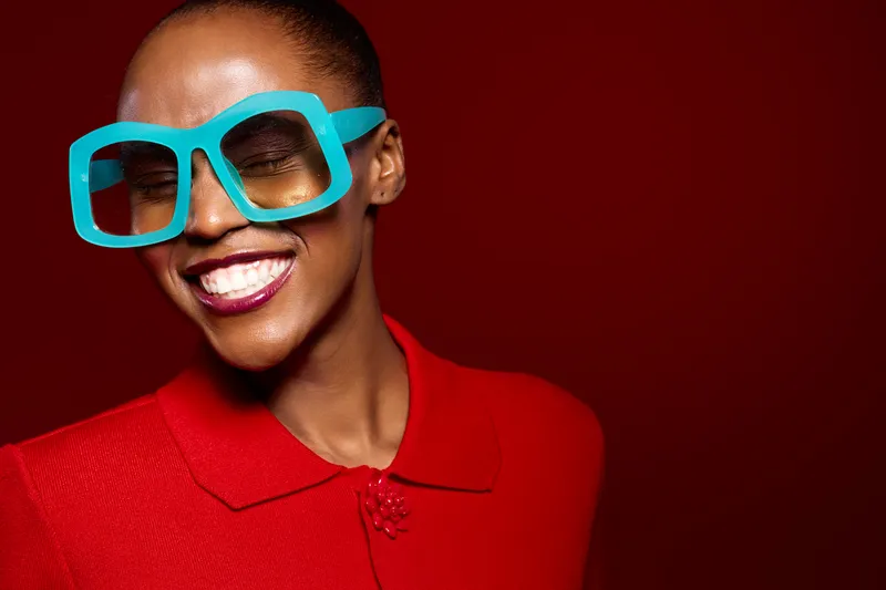 Smiling African American woman with dark skin wearing oversized blue glasses and red outfit poses against red background. Concept of fashion, style, joy, and vibrant personality.