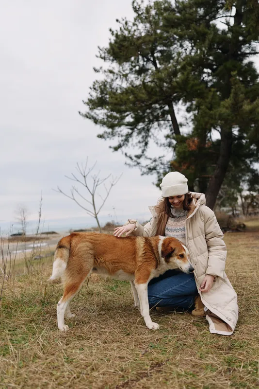 Dog and woman outdoors in a park, kneeling beside a friendly canine, sharing a quiet moment by a lakeside with trees in the background.
