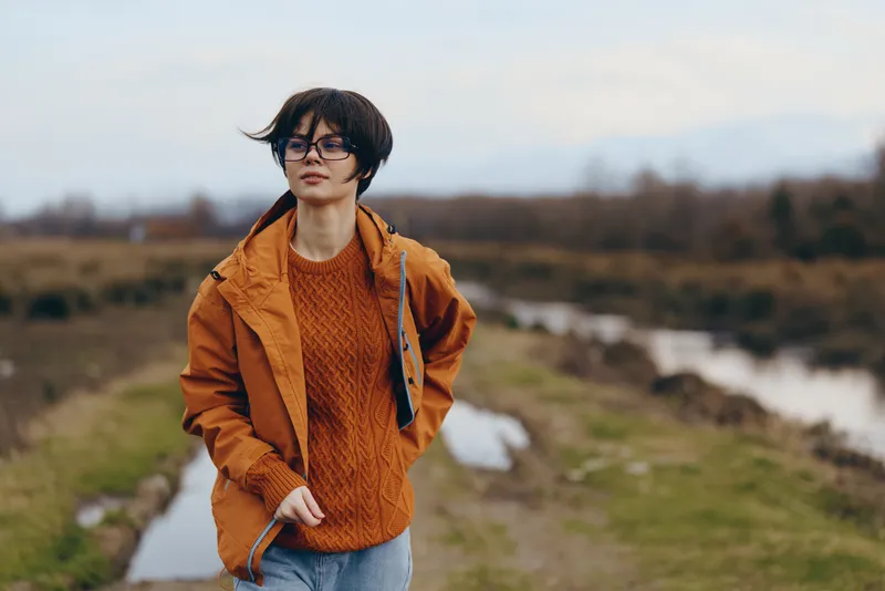 Woman lifestyle outdoors river landscape jacket sweater glasses countryside field by river orange knit sweater glasses natural morning light soft focus calm active daily walk in nature
