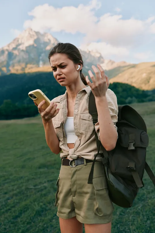 A young woman with a backpack stands in a sunlit grassy valley, scrolling her phone and frowning, conveying frustration and uncertainty during a hiking moment outdoors.