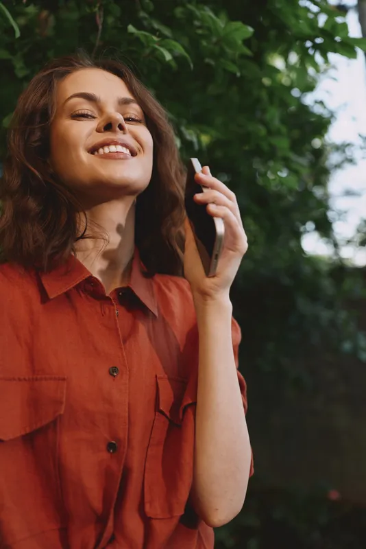 A smiling woman speaks on a phone outdoors, wearing a red shirt, as sunlight filters through trees and greenery, capturing a casual, joyful lifestyle moment