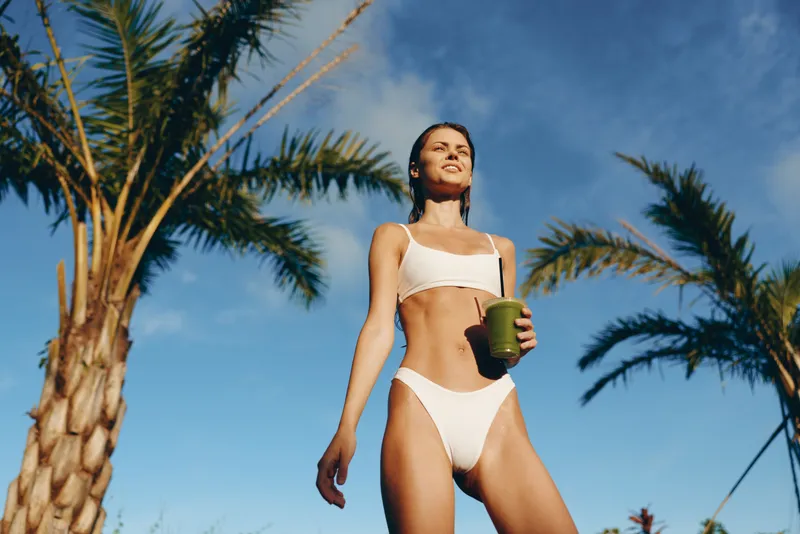 Young woman in white bikini holds green smoothie cup stands outdoors near palm trees under clear blue sky enjoying healthy lifestyle and sunny warm weather.
