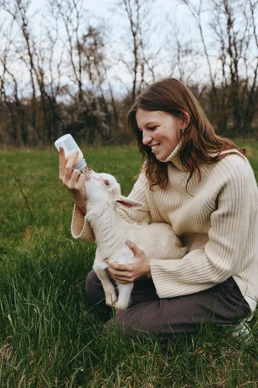 A woman in a knitted sweater bottle feeds a young goat outdoors, amid grass and trees. A tender moment of caregiving, farm lifestyle, and animal bonding in a natural setting today.