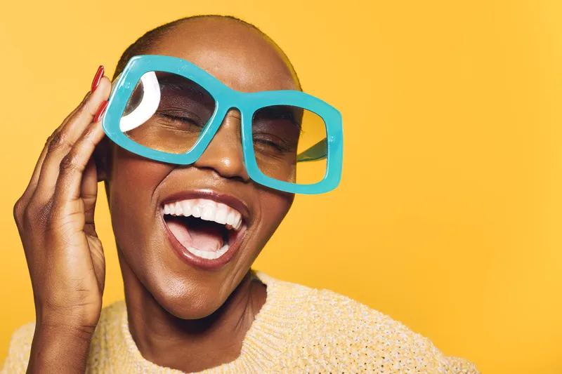 Happy dark skinned African woman with shaved head laughs joyfully wearing oversized blue sunglasses on yellow background. Concept of fashion, summer, confidence, and positive mood.