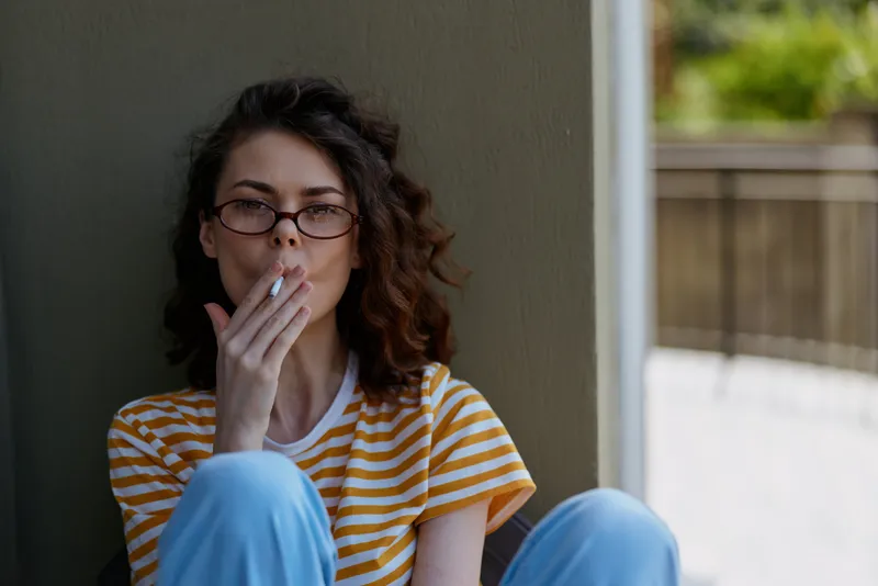 Woman with glasses sits indoors by a wall, wearing a striped yellow and white shirt and blue pants, smoking a cigarette in a quiet candid moment at home.