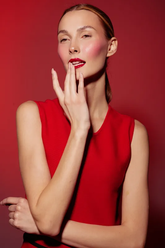 Young woman in red sleeveless top poses with fingers near lips against red background. Concept of fashion, beauty, makeup, and confident female model portrait.