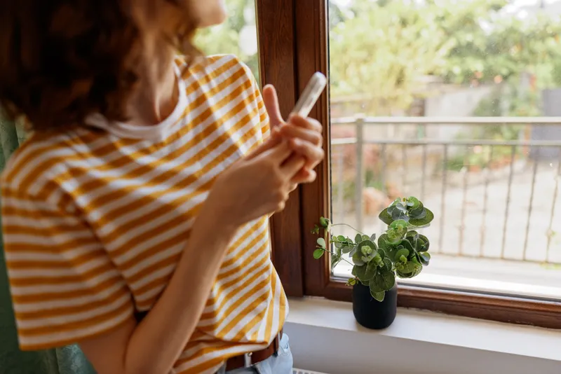 Woman at home in a striped shirt checks her smartphone by a sunny window, with a potted plant nearby, conveying a casual lifestyle and peaceful indoor moment. The scene feels intimate and relaxed.