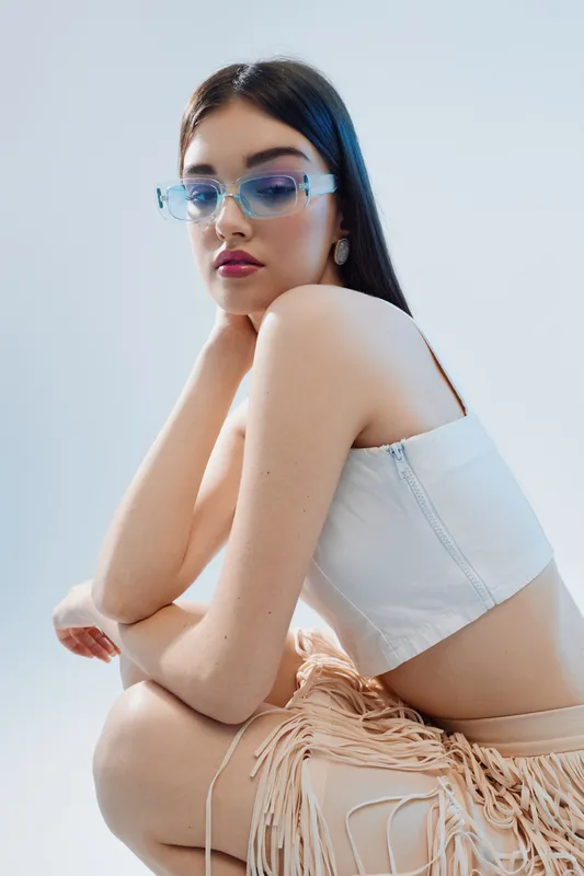 Young woman beauty fashion model wearing stylish sunglasses and white crop top posing against light background. Concept for modern style and elegance.