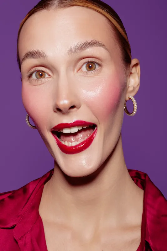 Close-up of a smiling woman with red lipstick and hoop earrings on purple background. Concept of beauty, makeup, fashion, and joyful expression in studio lighting.
