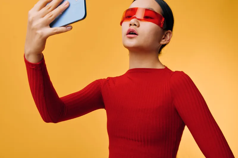 Young woman in red sweater and futuristic sunglasses taking selfie with smartphone on orange background. Fashion portrait with modern style, confident pose and digital technology concept.