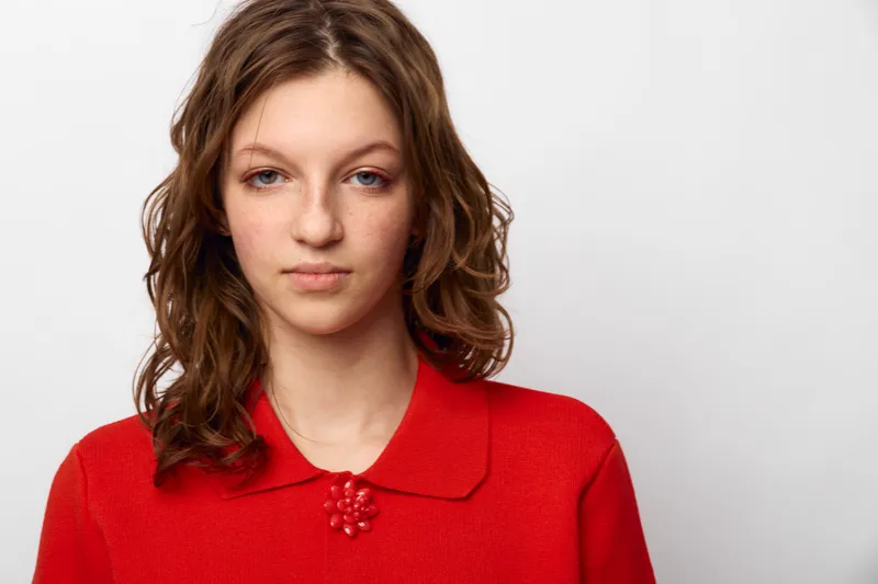 Young woman with wavy brown hair wearing red sweater looks calmly at camera on white background. Portrait of casual style, natural beauty, and confident female model.