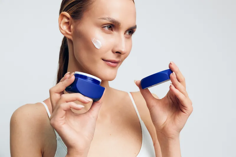 A young woman in a white tank top applies cream from blue jars to her cheek in a bright studio, illustrating a skincare routine and self care during a calm beauty moment.