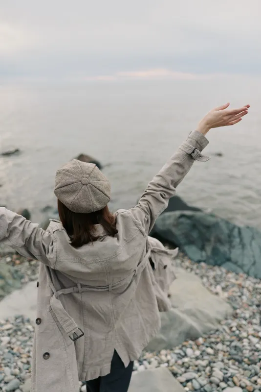Woman in beige coat and hat stands on rocky shore with arms raised facing calm sea. Concept of freedom, nature, travel, and peaceful outdoor moment by the water.