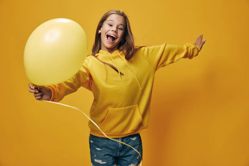 Happy girl in yellow hoodie holding balloon and smiling with excitement against orange background. Casual young woman celebrating with raised arm and joyful expression indoors.