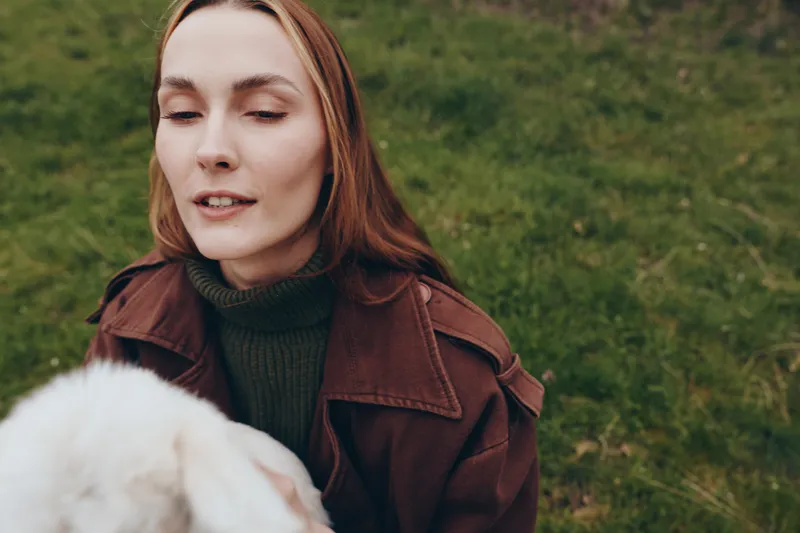Young woman with long hair wearing a brown jacket interacts with a white baby goat outdoors in a grassy area, capturing a natural and calm lifestyle moment.