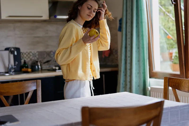 Bright woman in a yellow cardigan stands in a sunlit kitchen, holding a fruit and smiling softly, capturing a warm home moment, casual pose, natural colors, cozy atmosphere