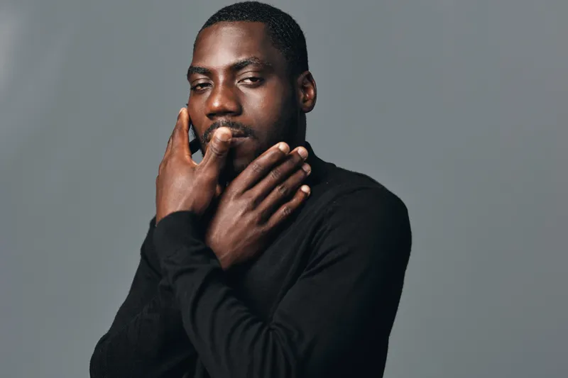 Creative visual designer style portrait of African American man with dark skin wearing black sweater posing with hands near face on gray background in studio lighting.