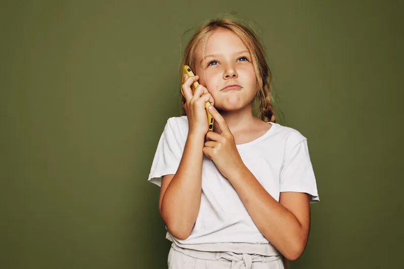 Young girl with blonde hair in a white t-shirt holds a yellow phone to her ear looking thoughtful on an olive green background.