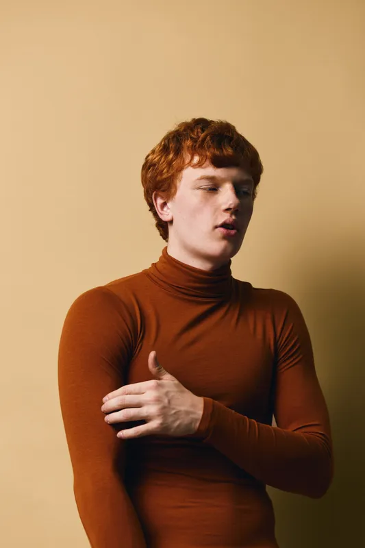Young man with red hair and pale skin wearing brown turtleneck sweater standing against beige background with calm expression and closed eyes.
