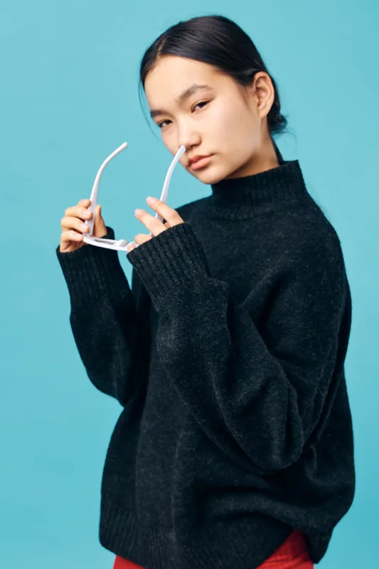 Young woman with black sweater putting on white sunglasses against blue background, posing with serious expression for fashion and style portrait photography in studio setting.