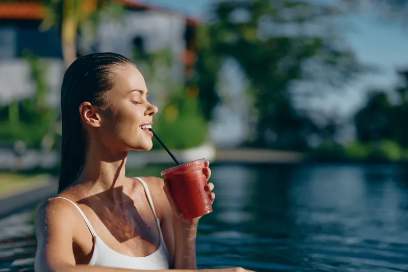 A woman relaxes poolside, sipping a red smoothie through a straw, wearing a white tank top. Bright sunshine highlights casual leisure by the water, conveying lifestyle, wellness, and refreshment.
