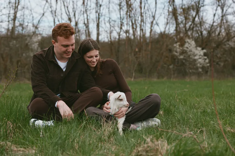 Joyful couple outdoors, sitting on grass with a playful goat kid nearby, warm sunlight, relaxed posture, gentle breeze, natural colors, lush surroundings, pastoral calm, intimate moment, countryside