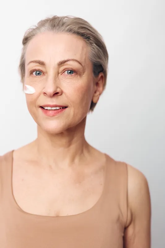 Mature woman with short blonde hair applies facial cream on cheek, smiling gently. Skin care concept with natural beauty, wellness, and self-care on white background studio portrait.