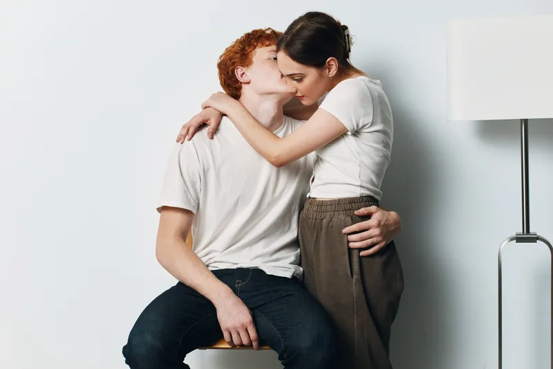 Couple sitting and hugging showing affection and love with casual clothes in minimal studio space. Young man and woman sharing intimate moment with gentle embrace and kiss on cheek.