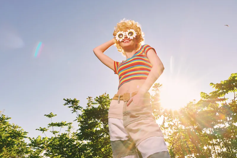 Cheerful woman outdoors, wearing striped top and floral sunglasses, enjoying sunny light, relaxed pose, retro film color vibe, lush greenery background, candid lifestyle moment